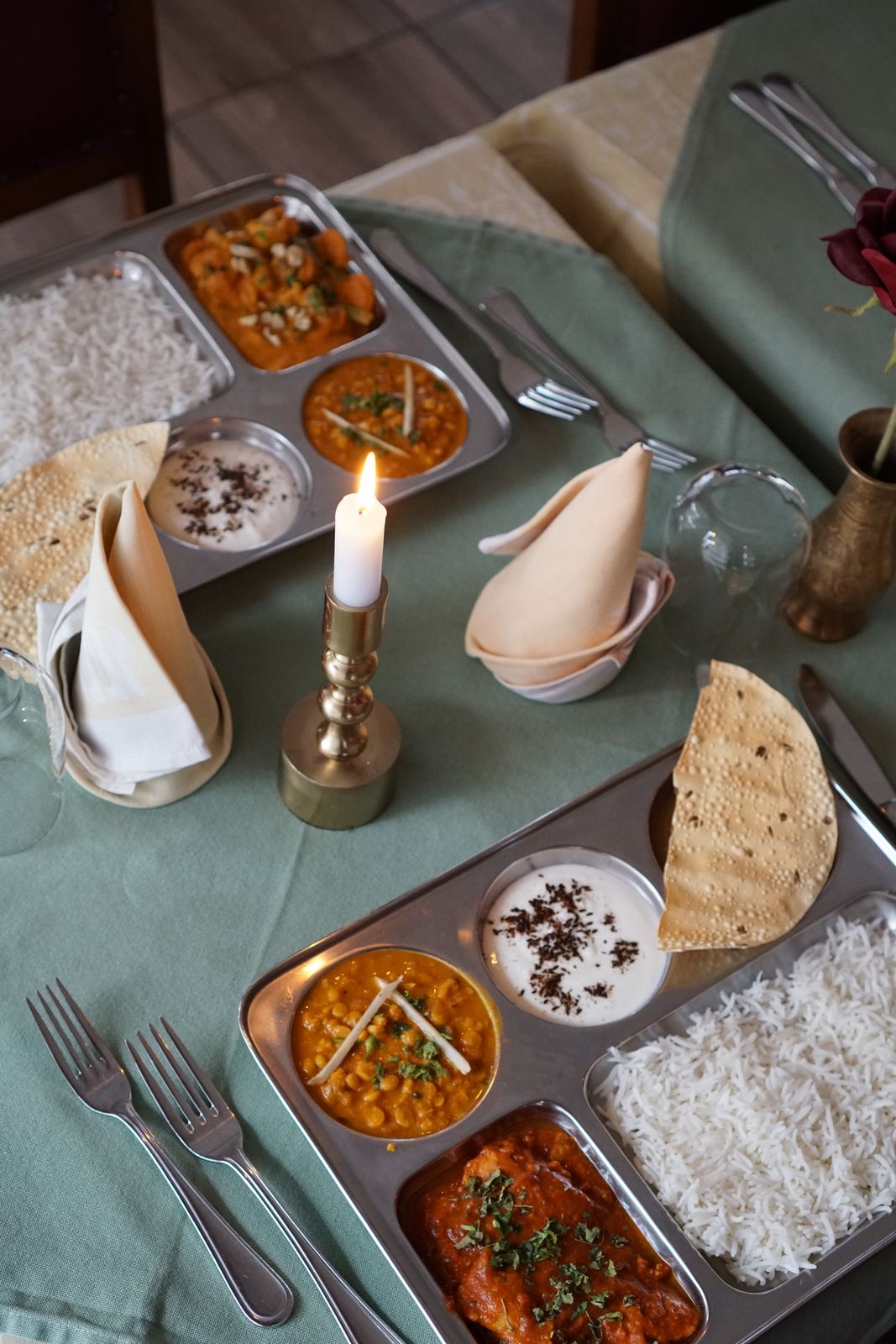 Zwei Thali-Platten mit Curry, Dal, Raita, Basmatireis und Papad auf einem gedeckten Tisch mit Kerze.